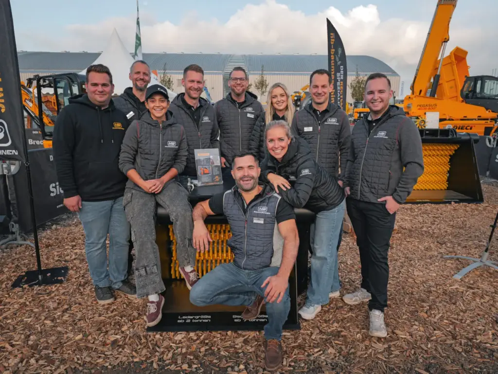 Mehrere Teammitglieder von NB Baumaschinen stehen auf einem Freigelände zwischen orangefarbenen Maschinen und Messeständen für ein Gruppenfoto.