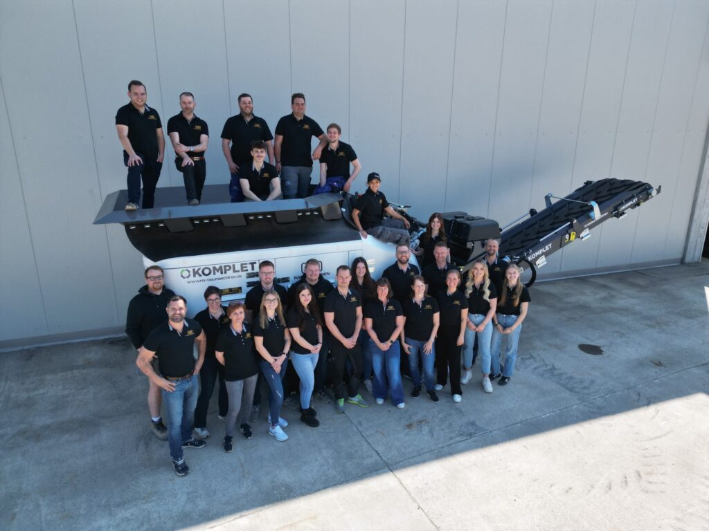 Vor einer hellen Wand posiert ein großes Team in schwarzen Poloshirts um eine schwarze KOMPLET Siebanlage mit ausfahrbarem Förderband.