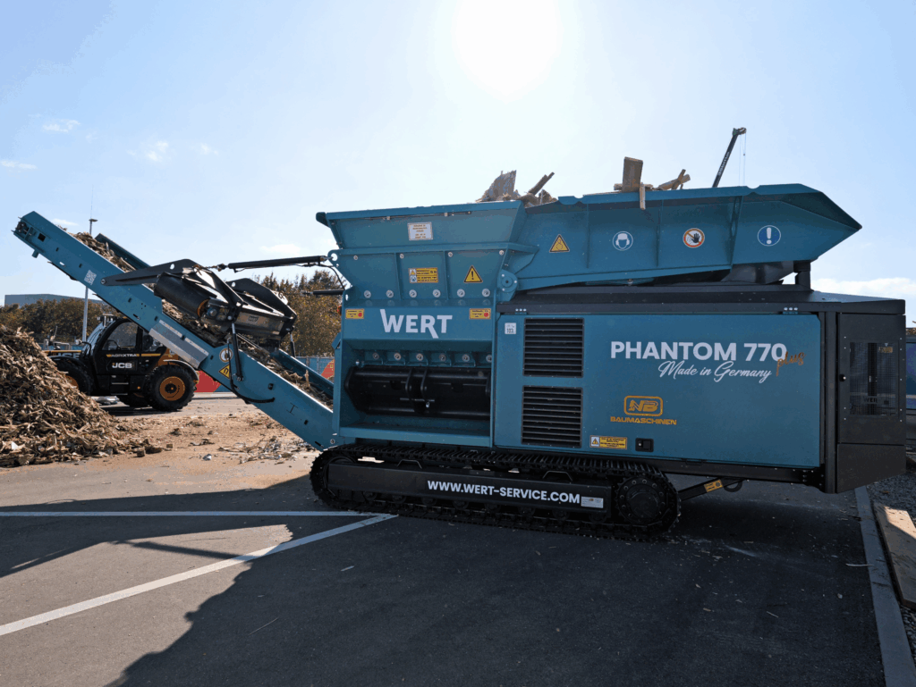 Unter blauem Himmel arbeitet der blaue WERT PHANTOM 770 plus von NB Baumaschinen neben einem Radlader, während Holzreste auf das Förderband laufen.