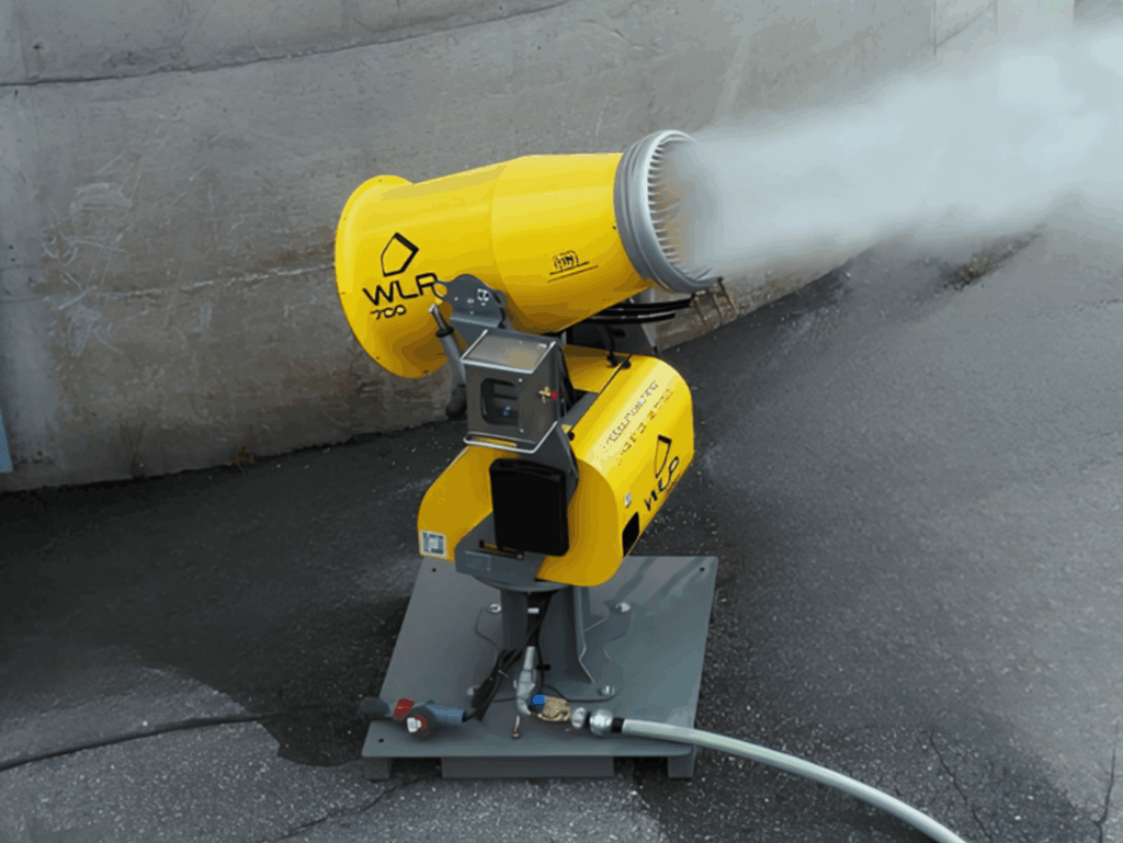Der gelbe WLP 700 zerstäubt dichten Wassernebel aus einer Düse, während er mit angeschlossenem Schlauch auf dunklem Asphalt vor einer Betonwand steht.