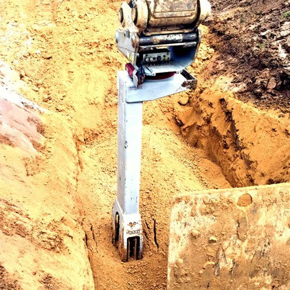 Ein Baggeraufsatz steht in einem schmalen, erdigen Graben, während daneben ein helles Metallteil und ein brauner Erdwall zu sehen sind.