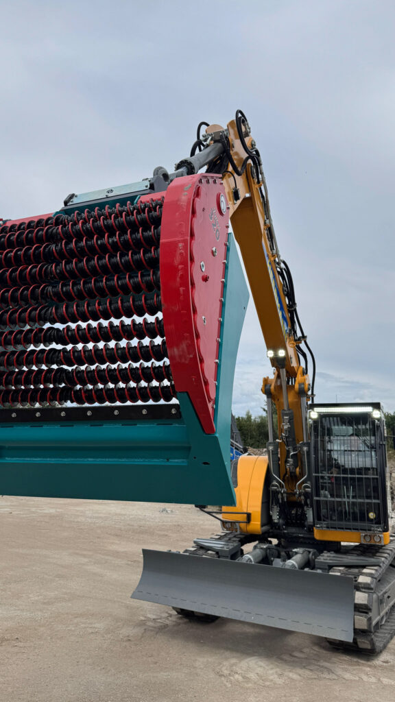 Ein gelber Raupenbagger mit langem Ausleger trägt einen großen roten Sieblöffel auf einem Betonplatz bei einem Recyclingbetrieb unter grauem Himmel.