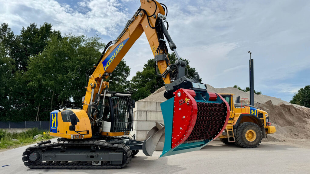 Ein gelber Kettenbagger mit großem Sieblöffel arbeitet neben einem Radlader vor Sandhaufen, Bäumen und bewölktem Himmel auf einem Schotterplatz.