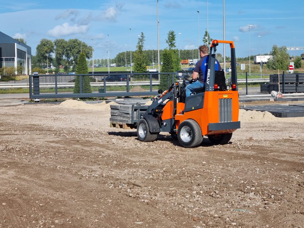 Ein orange-schwarzer RELLY-Radlader fährt über einen Schotterplatz, während der Fahrer Material an einem Baugelände mit Zaun und Sandhaufen bewegt.