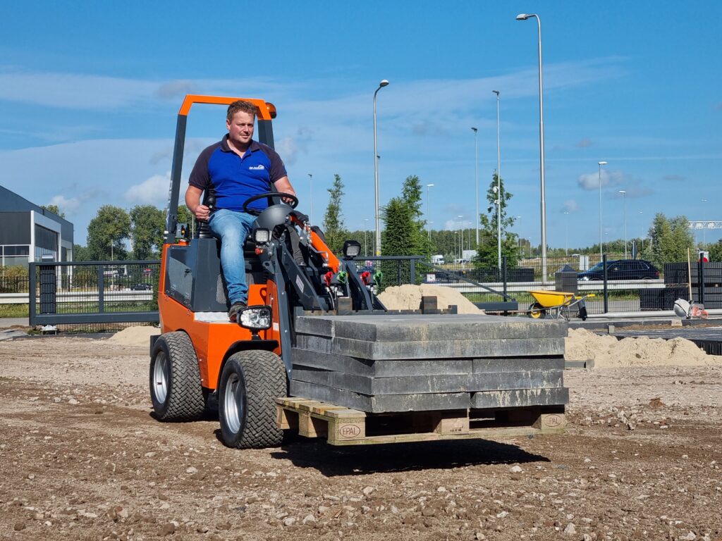 Ein Mann fährt einen orange-schwarzen Hoflader mit schweren Betonplatten auf einer Palettenladung über eine Baustelle im Freien bei blauem Himmel.