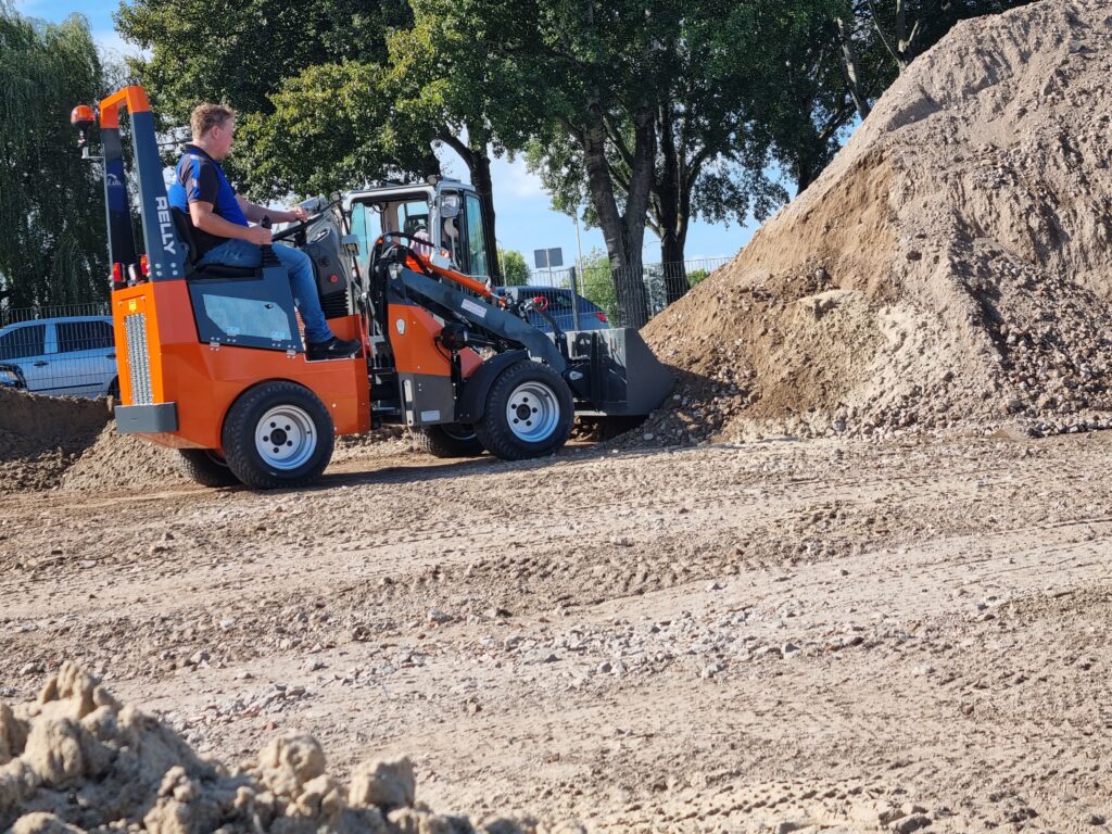 Ein orange-schwarzer Radlader schiebt auf einem sandigen Platz Material gegen einen großen Erdhaufen, während ein Mann am Steuer sitzt im Freien.