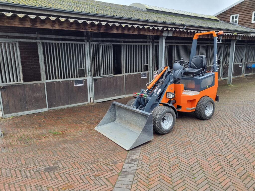 Ein orange-grauer Radlader steht vor Pferdeställen auf einem gepflasterten Hof, mit großer Schaufel, Kabine, breiten Reifen und Warnleuchte.