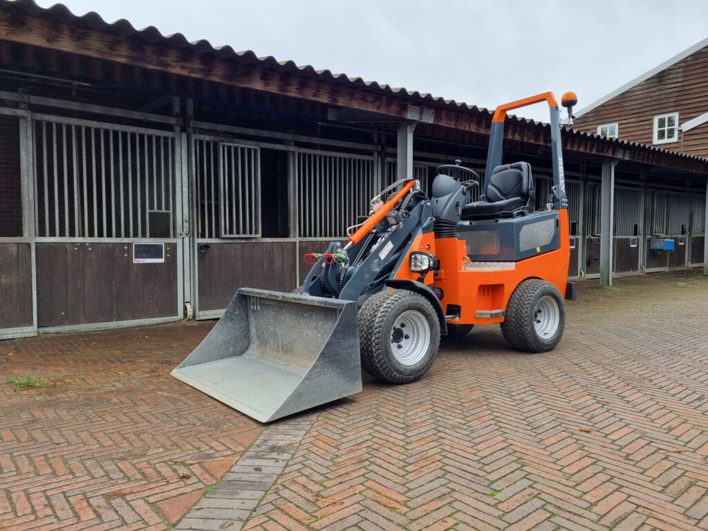 Ein orange-grauer Radlader mit großer Schaufel steht auf einem Pflasterhof vor dunklen Stallgebäuden mit Holzfassade und Gittertoren.