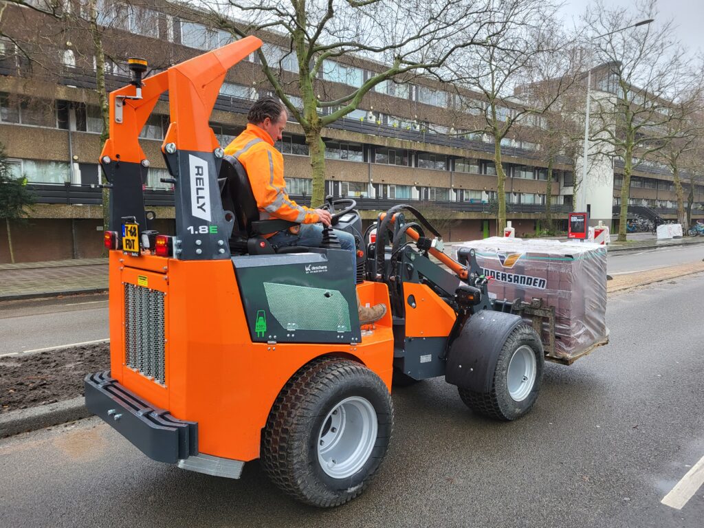 Ein orangefarbener RELLY 1.8E mit Fahrer transportiert auf einer nassen Straße eine palettierte Last vor grauen Wohnhäusern und kahlen Bäumen.