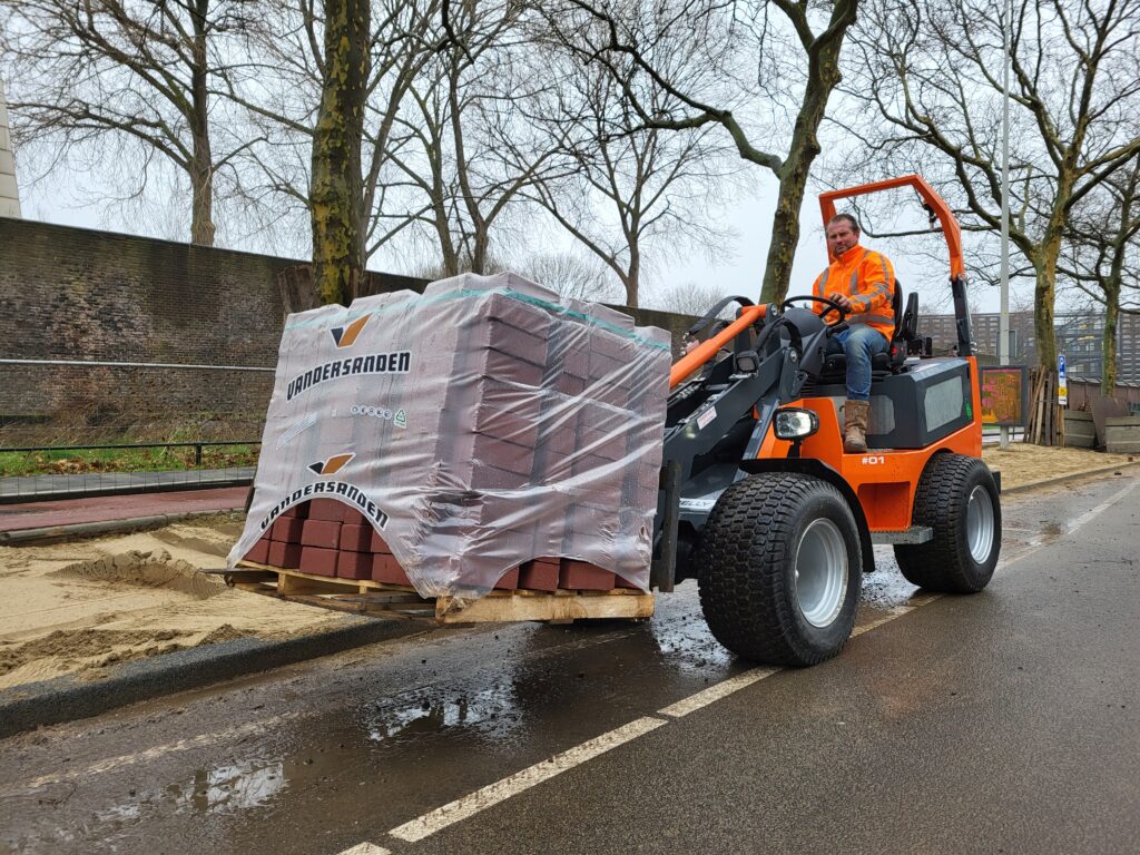 Ein Mann in orangefarbener Arbeitskleidung fährt einen orange-schwarzen Radlader mit palettierter Ziegelsteinladung an einer nassen Straße entlang.