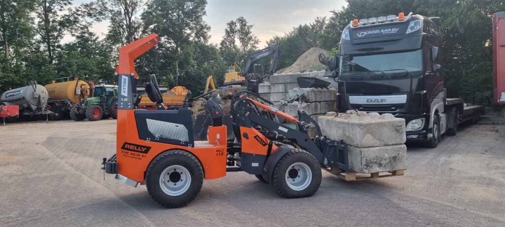 Ein orange-schwarzer RELLY-Radlader hebt große Betonblöcke neben einem schwarzen DAF-Lkw auf einem Betriebshof mit weiteren Baumaschinen.