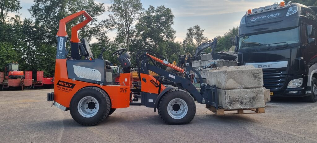Ein orange-schwarzer RELLY-Lader hebt große Betonblöcke neben einem DAF-Lkw auf einem Recyclinghof mit Bäumen im Hintergrund bei Tageslicht.