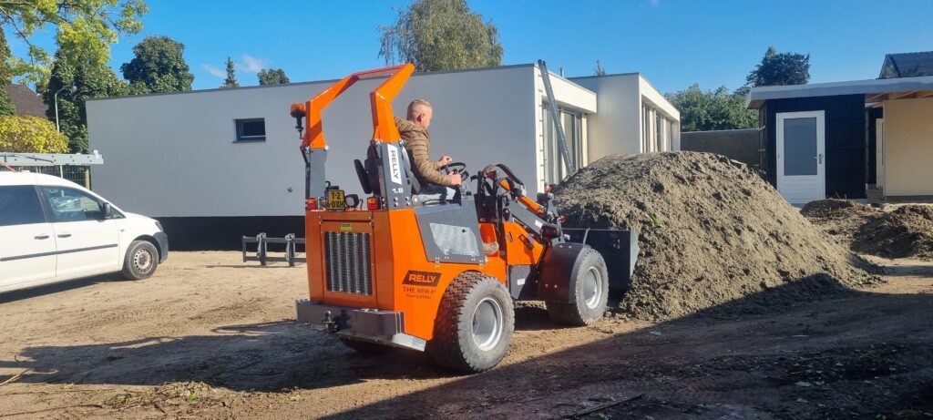 Ein orangefarbener Rely-Lader mit angehobener Schaufel bewegt auf einer Baustelle neben einem Sandhaufen Erde, während ein Mann die Maschine steuert.