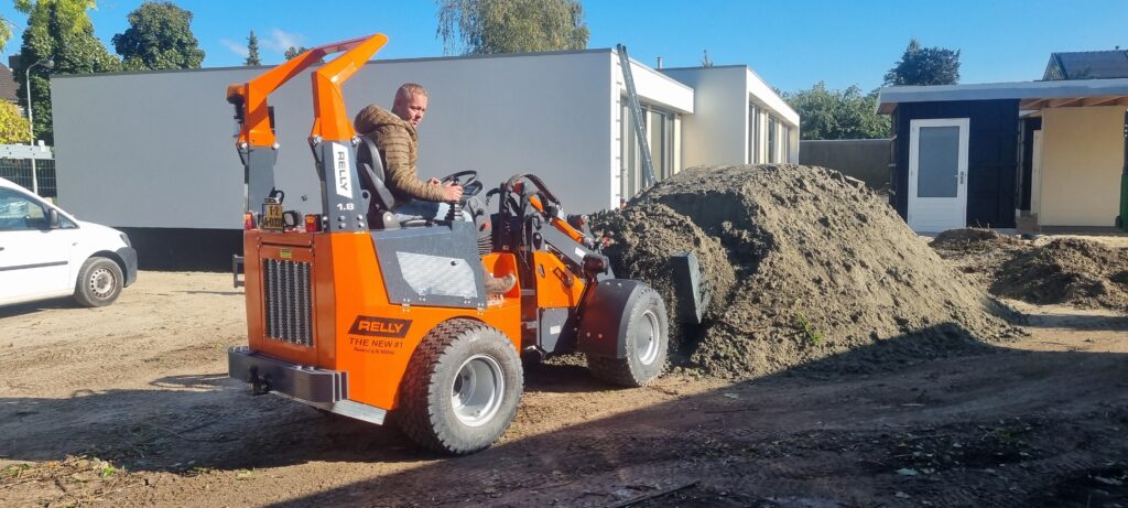 Ein orangefarbener Rely-Radlader schiebt auf einem sonnigen Betriebshof einen großen Sandhaufen, während der Fahrer die offene Maschine steuert.