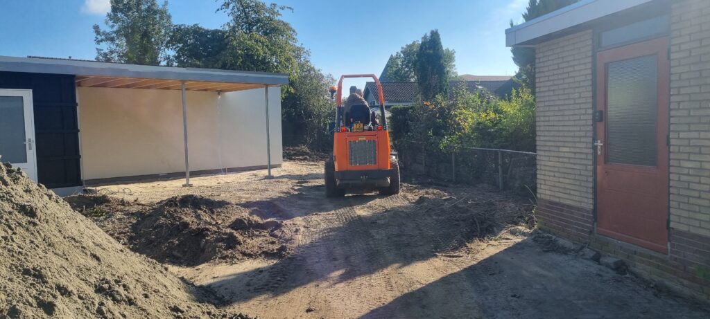 Ein orangefarbener Kompaktlader fährt über ein sandiges Baugrundstück zwischen zwei Gebäuden, während im Hintergrund Bäume und Hecken stehen.