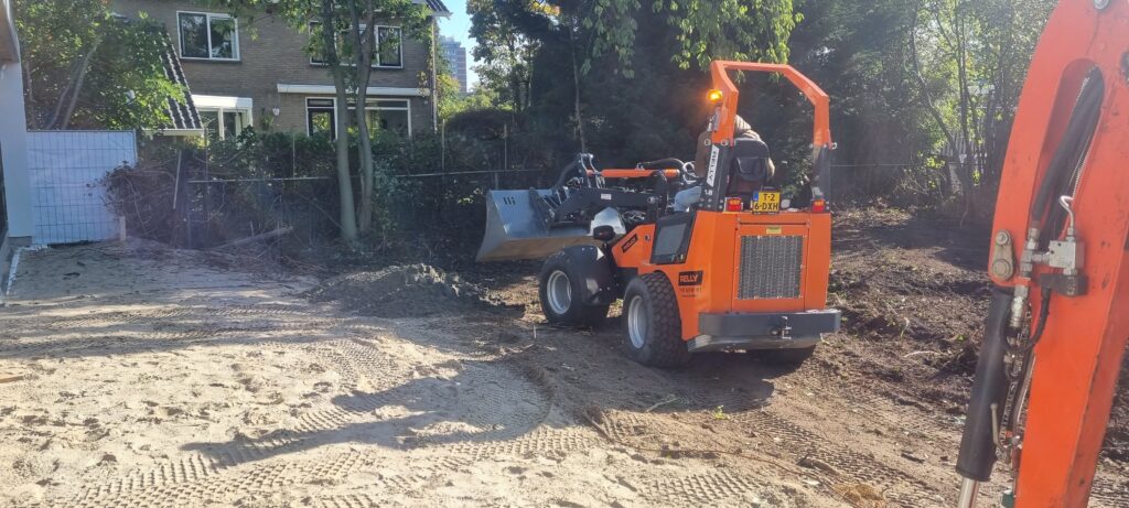 Ein orangefarbener Radlader mit Schaufel fährt auf einer sandigen Baustelle vor einem Wohnhaus, links Brachfläche und rechts ein Baggerarm.