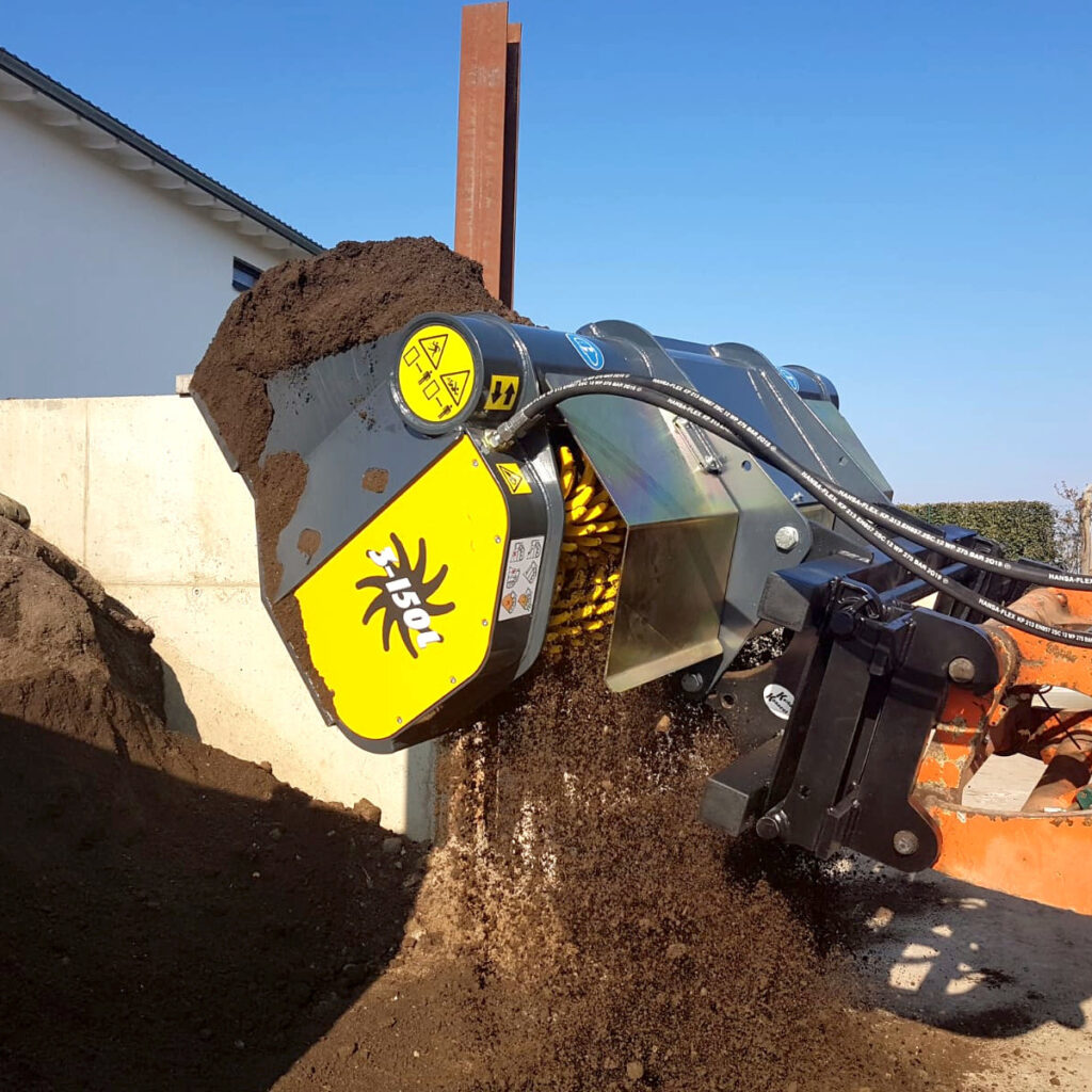 Ein gelber und schwarzer Sieblöffel mit der Aufschrift 150L trennt Erde auf einer Baustelle, während feines Material nach unten rieselt.