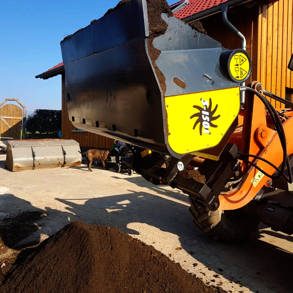 Ein großer schwarz-gelber Recycling-Baumaschinenaufsatz hebt Erde vor einem Hof, während ein Hund daneben steht und ein weiteres Anbaugerät liegt.
