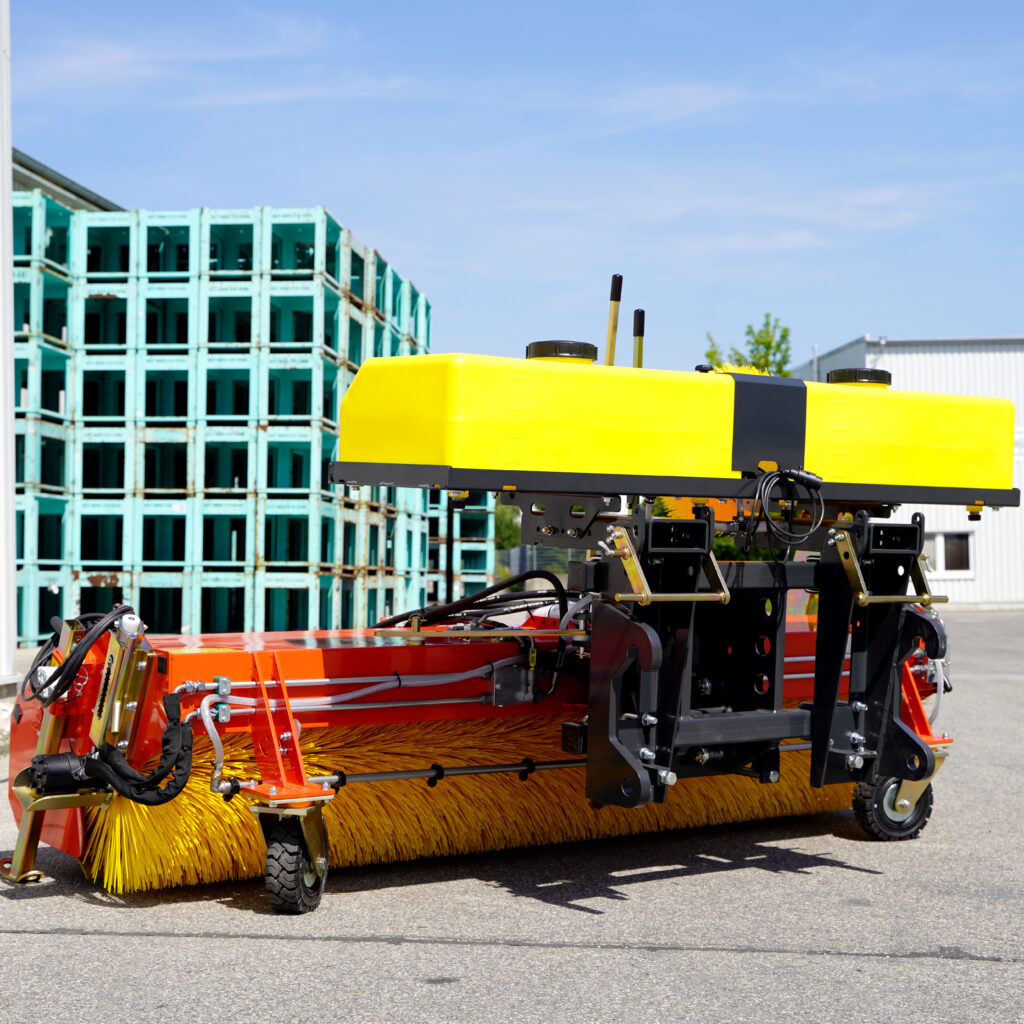 Ein gelb-orangefarbener Adler Kehrmaschine K600-3 mit Bürstenwalze steht vor einem Industriehof und zeigt robuste Anbautechnik.