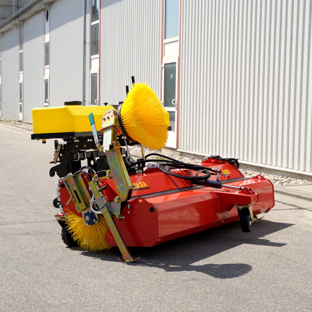 Vor einem hellen Industriebau steht eine rote Adler-Kehrmaschine mit gelbem Bürstenkranz, Hydraulikleitungen und kompaktem Aufbau auf Asphalt.