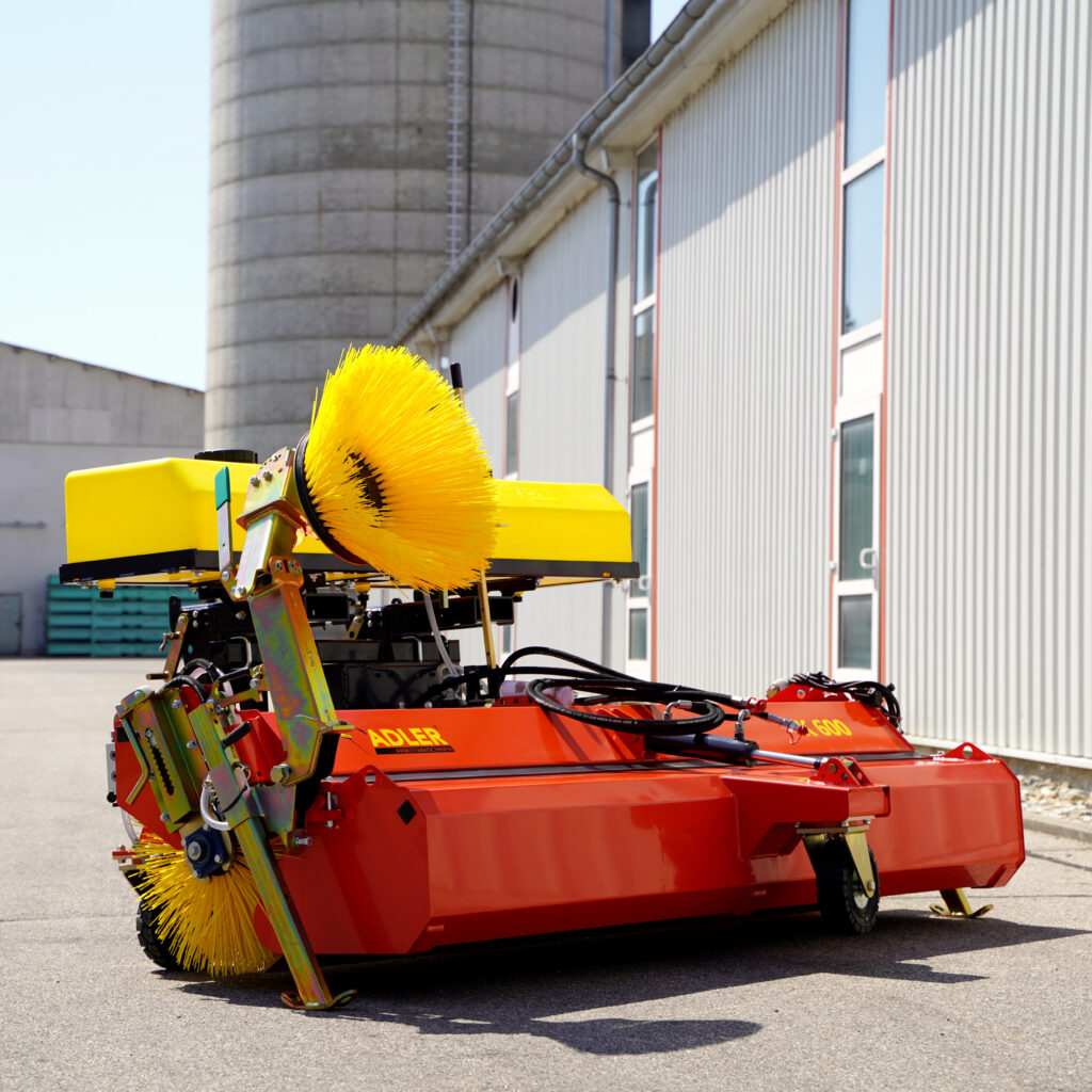 Vor einer Industriehalle steht eine rote Adler-Kehrmaschine mit gelbem Bürstenelement, sichtbaren Hydraulikschläuchen und großen Silos im Hintergrund.