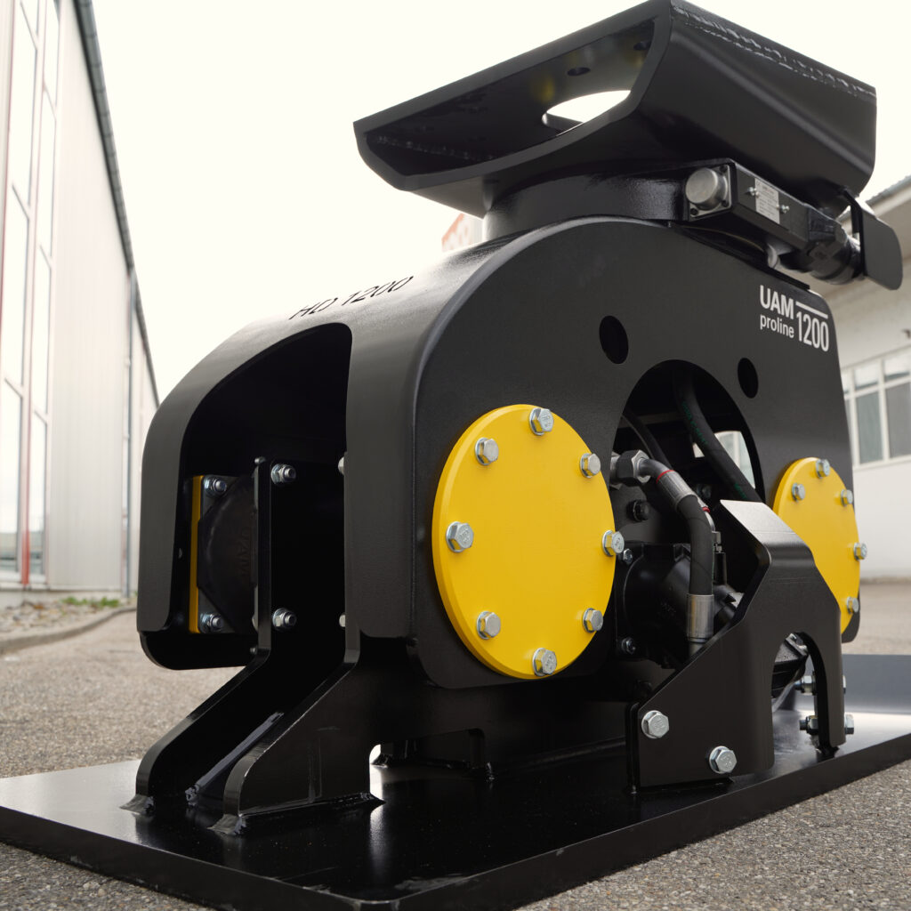 Der schwarze Anbauverdichter UAM proline 1200 mit gelben Abdeckungen und massiver Stahlbasis steht vor einem Gebäude auf Pflaster.