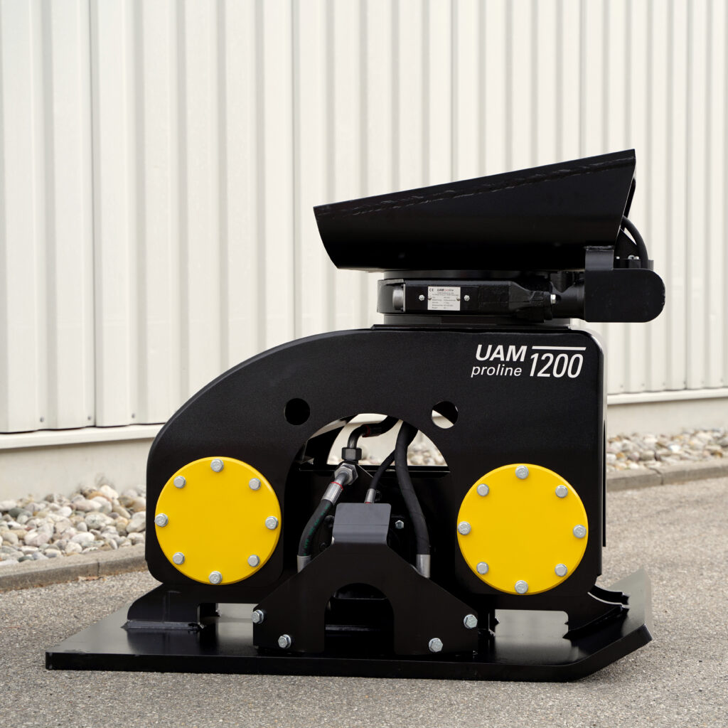 Ein schwarzer Anbauverdichter mit gelben Rundplatten und der Aufschrift UAM proline 1200 steht auf Asphalt vor einer hellen Wellblechwand.