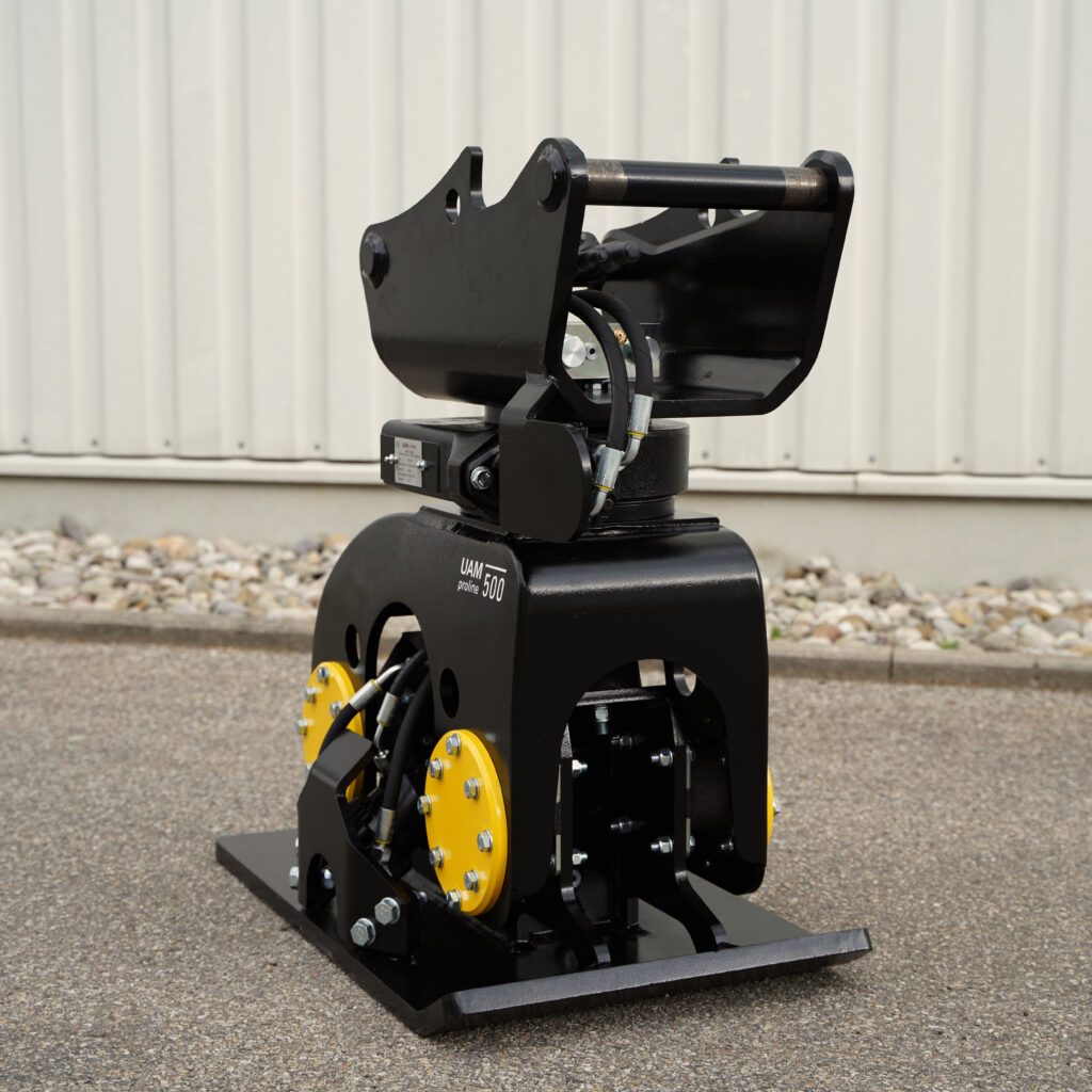 Ein schwarzer Anbauverdichter mit gelben Seitenteilen, Hydraulikschläuchen und massiver Stahlkonstruktion steht vor einer hellen Wand auf Asphalt.