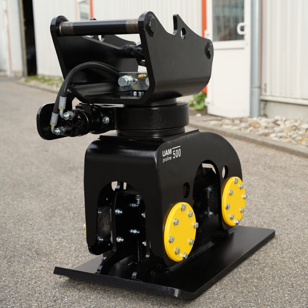 Schwarzer Anbauverdichter mit massiven Stahlarmen, Hydraulikschlauch und gelben Abdeckungen steht vor einer Industriehalle auf Asphalt.