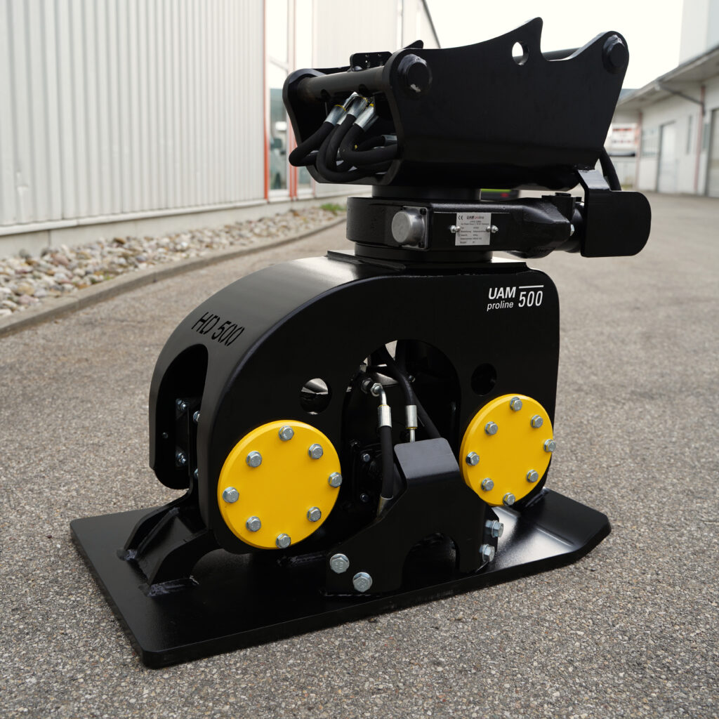 Schwarzer Anbauverdichter HD 500 von UAM proline mit gelben Abdeckungen und Hydraulikschläuchen steht vor einer Industriehalle auf Asphalt.