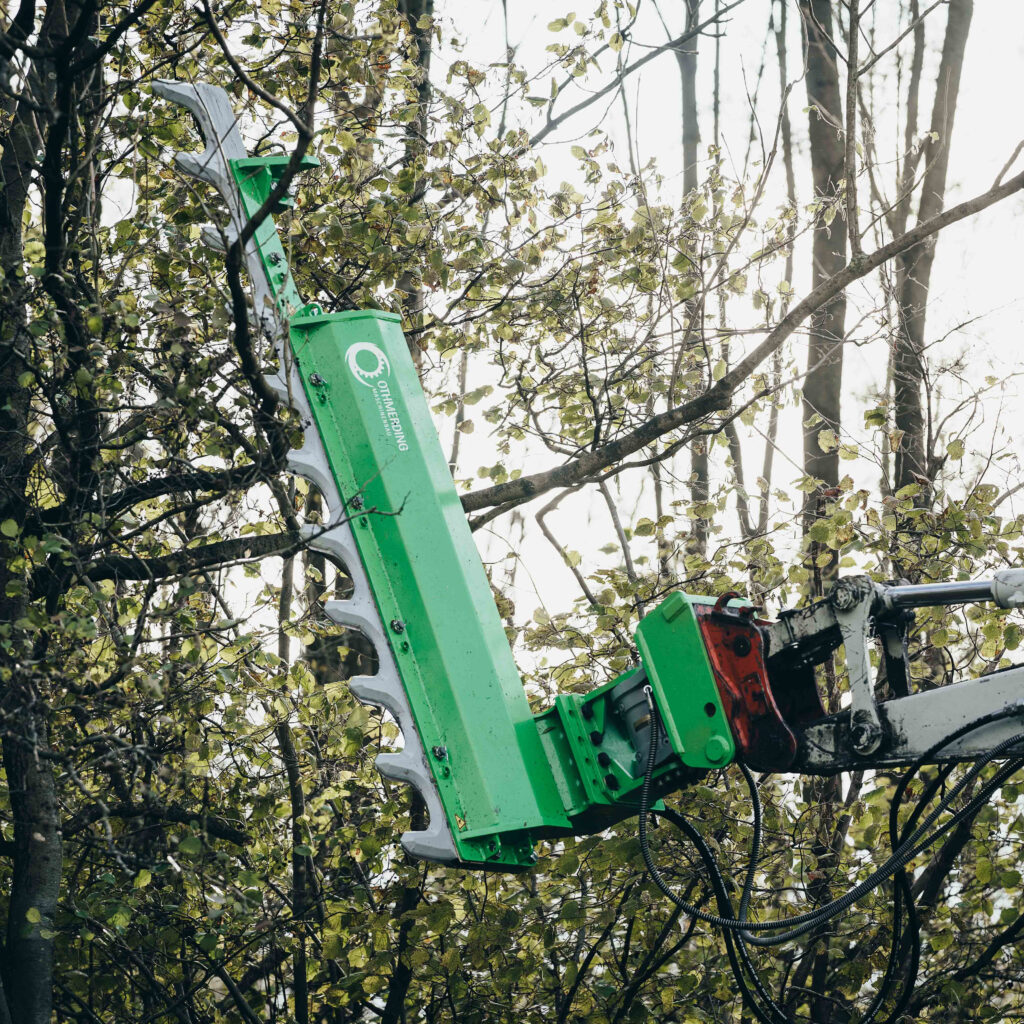 Ein grüner OTHMERDING-Ast- und Heckenschneider greift zwischen belaubte Äste, während Hydraulikschläuche und Stahlteile im Vordergrund stehen.