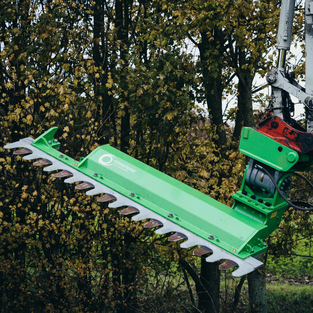 Eine grüne Othmerding AHS 240 Ast- und Heckenschere arbeitet zwischen dichtem Laub; rechts sind Hydraulikarm und Schneidbalken zu sehen im Baumwerk.