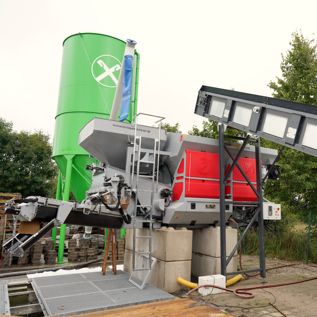 Eine grüne BETONstation S430-1 mit großem Siloturm, grauen Modulen und rotem Gehäuse steht im Freien zwischen Bäumen und Betonfundamenten.