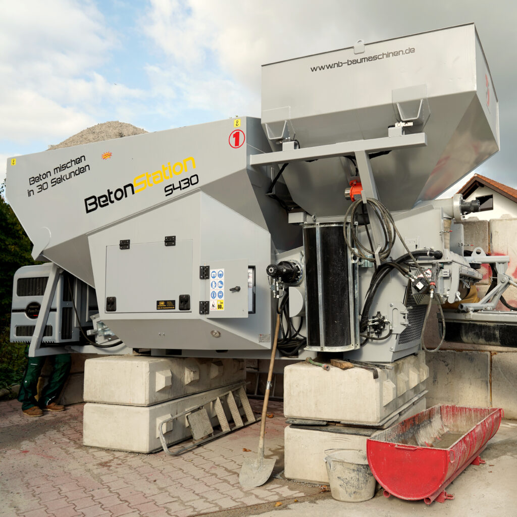 Eine graue BETONstation S430 mit großen Trichtern, Schläuchen und Hydraulikkomponenten steht auf Betonblöcken unter einem hellen Himmel.
