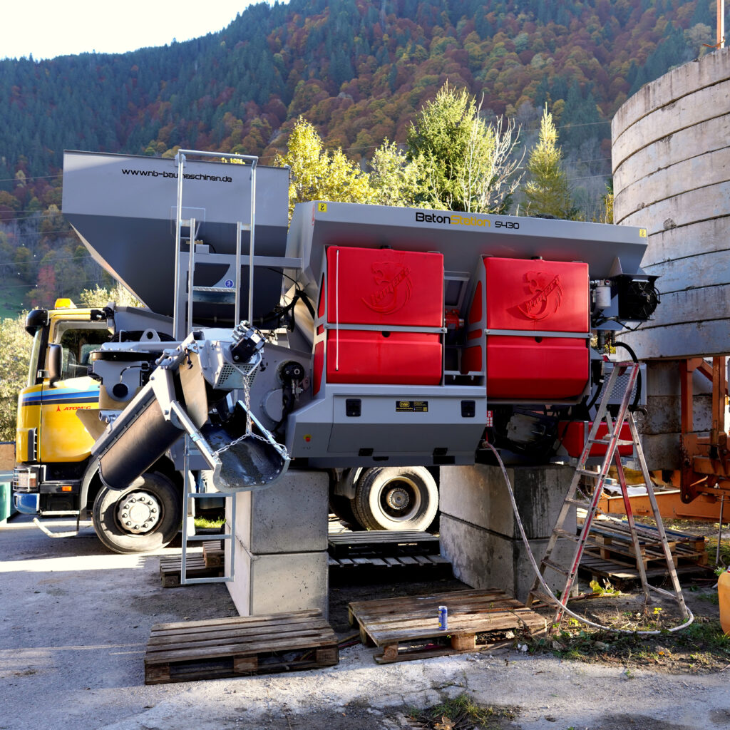 Die BetonStation S430 steht mit grauen Behältern und roten Abdeckungen auf einem Lkw vor bewaldetem Berghang und silbernem Silo.