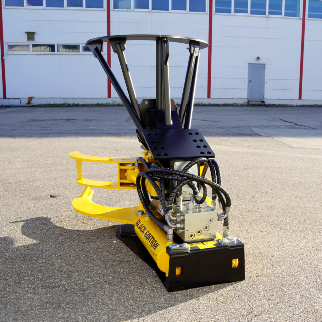 Eine gelb-schwarze Baumschere mit Hydraulikschläuchen und massiven Stahlarmen steht vor einer Industriehalle auf Asphalt.
