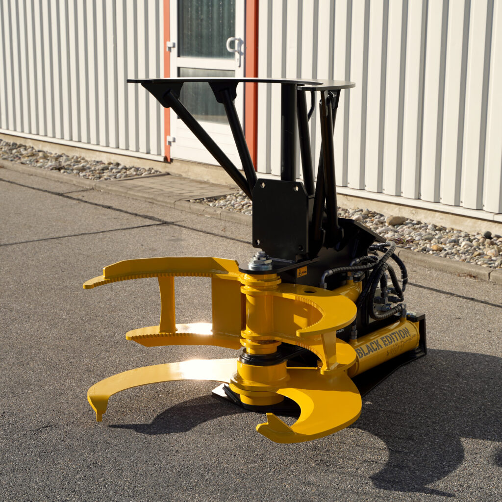 Schwarze und gelbe Baumschere mit hydraulischen Schläuchen steht vor einer hellen Metallfassade auf Asphalt im Sonnenlicht neben einem Eingangstor.