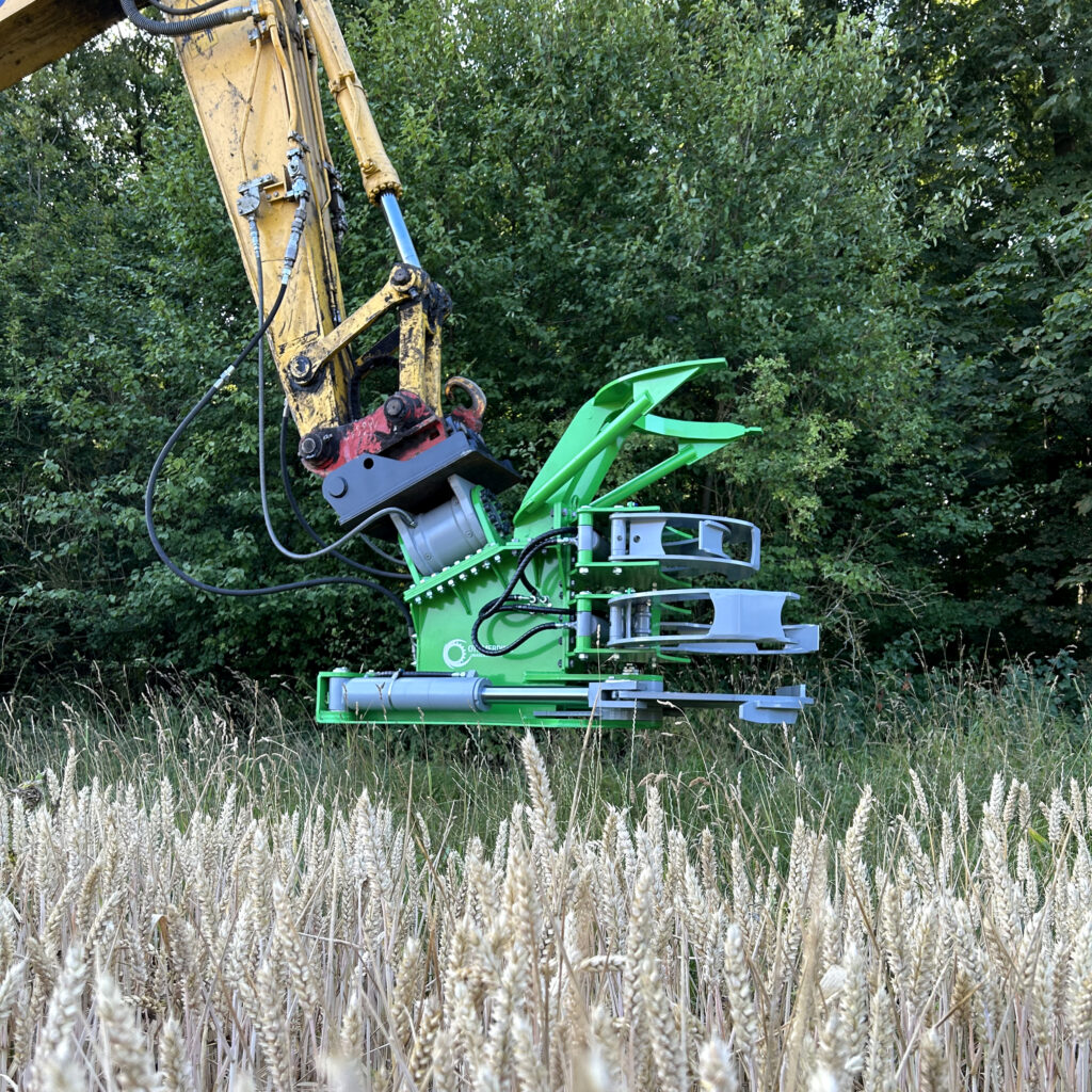 Ein gelber Baggerarm hält eine leuchtend grüne Baumschere am Rand einer Wiese, davor steht hohes Gras vor dichtem Wald im Sommerlicht.