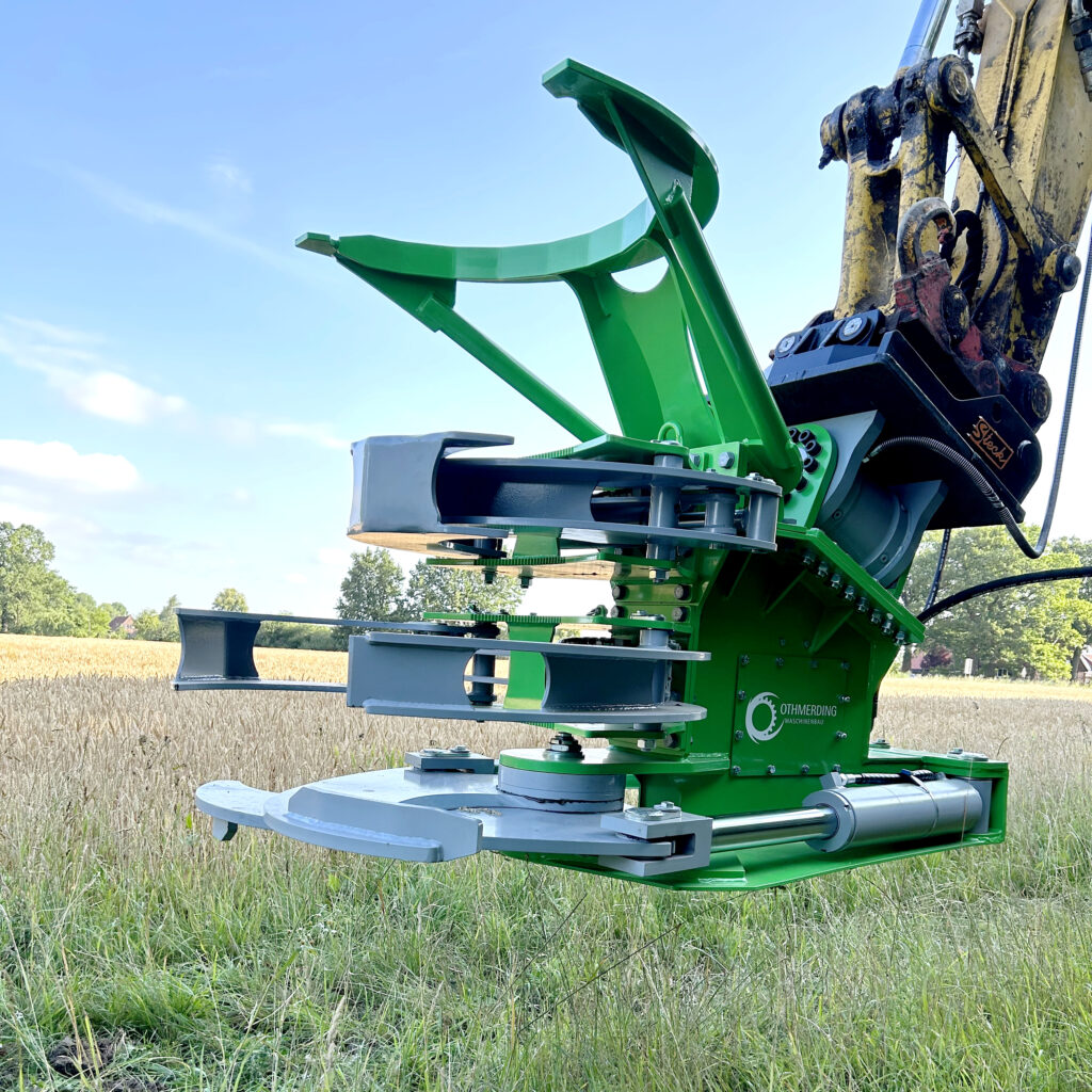 Eine grüne Baumschere mit grauen Schneidarmen hängt an einem gelben Baggerarm über einem Getreidefeld unter blauem Sommerhimmel und Wiesen.