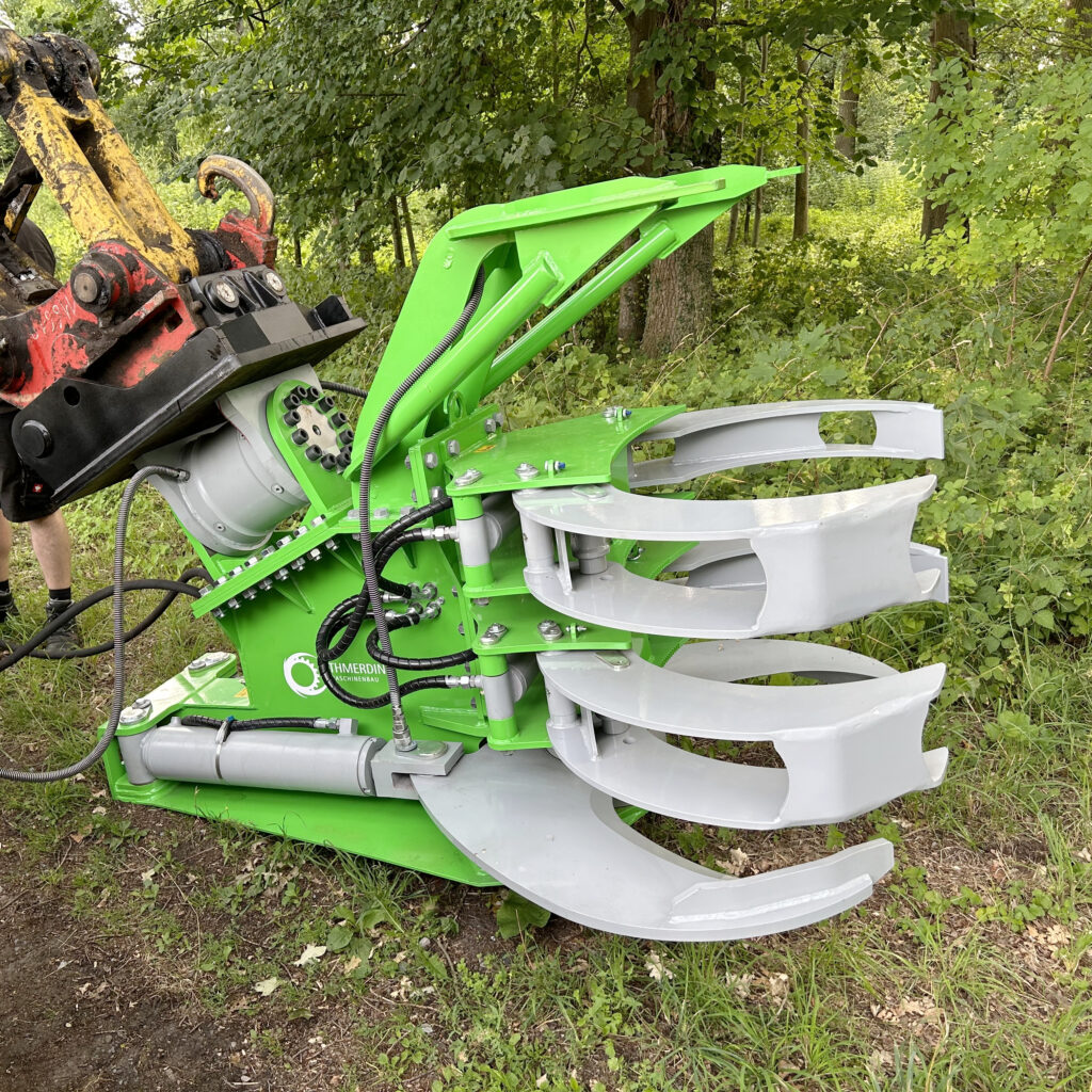 An einem grünen Baumgreifer mit silbernen Greifarmen hängt ein Baggerarm im Wald; Schläuche und Hydraulikzylinder sind gut sichtbar.
