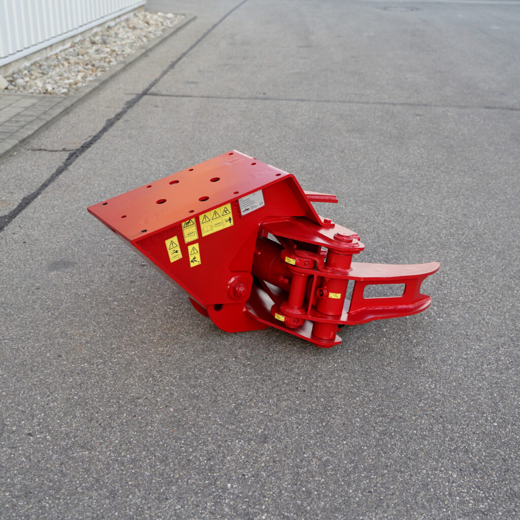 Ein rotes, hydraulisches Anbaugerät mit gelben Warnaufklebern liegt auf Asphalt vor einem Industriegebäude, mit breiter Aufnahme und massiven Scherenarmen.