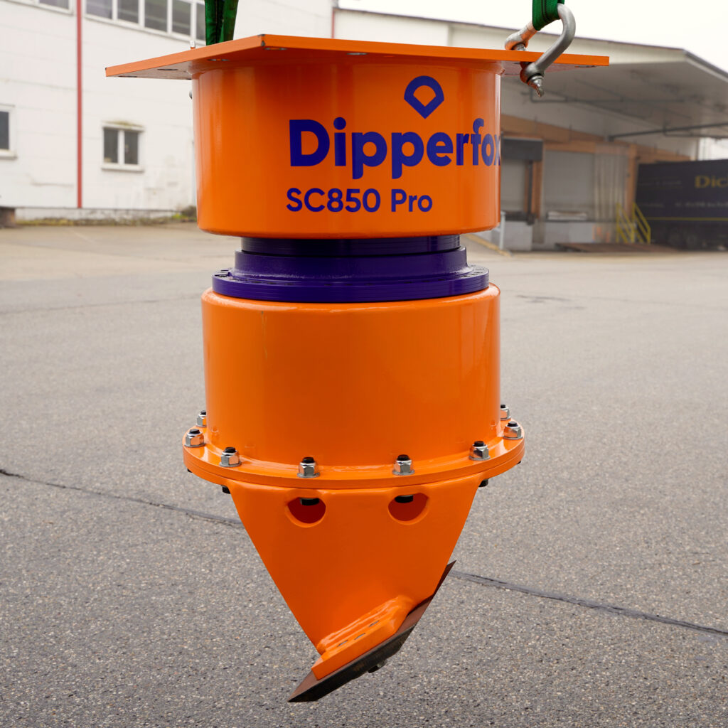 Vor einer Industriehalle hängt ein orange-violetter Baumstumpffräser SC850 Pro im Freien, mit sichtbaren Bolzen, Aufhängung und spitzem Fräskegel.