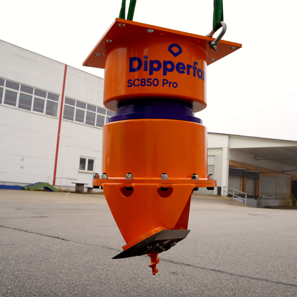 An einem Kran hängt ein orangefarbener Dipperfox SC850 Pro mit Bohrspitze vor einer Industriehalle; grüne Hebebänder und blaue Schrift sind sichtbar.