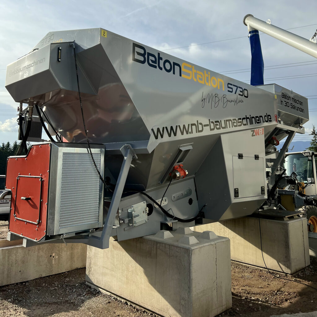 Eine graue Betonmischstation S730 von NB Baumaschinen steht auf Betonfundamenten, daneben sind rote und blaue Maschinenteile im Freien zu sehen.