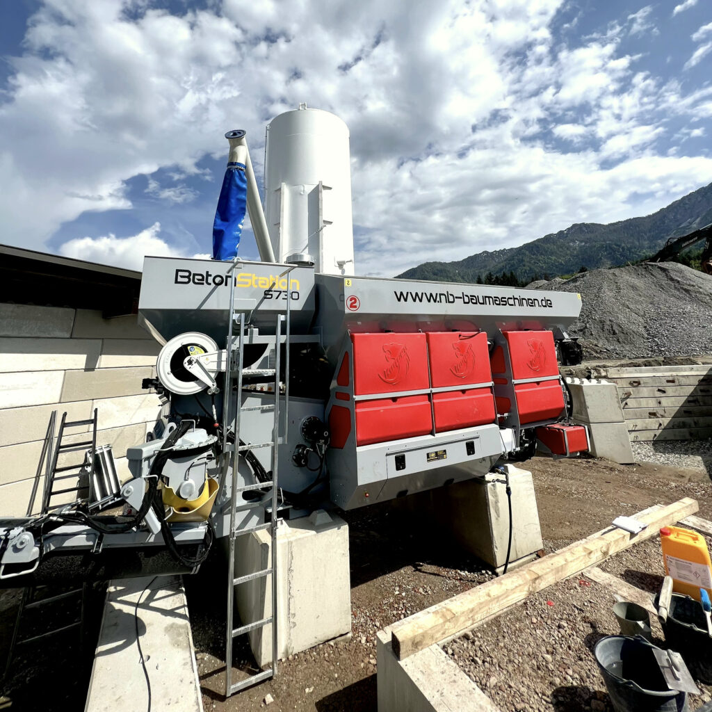 Eine weiße BetonStation S730 mit roten Wartungsklappen steht in bergiger Landschaft unter wolkigem Himmel, umgeben von Schotter und Baggern.