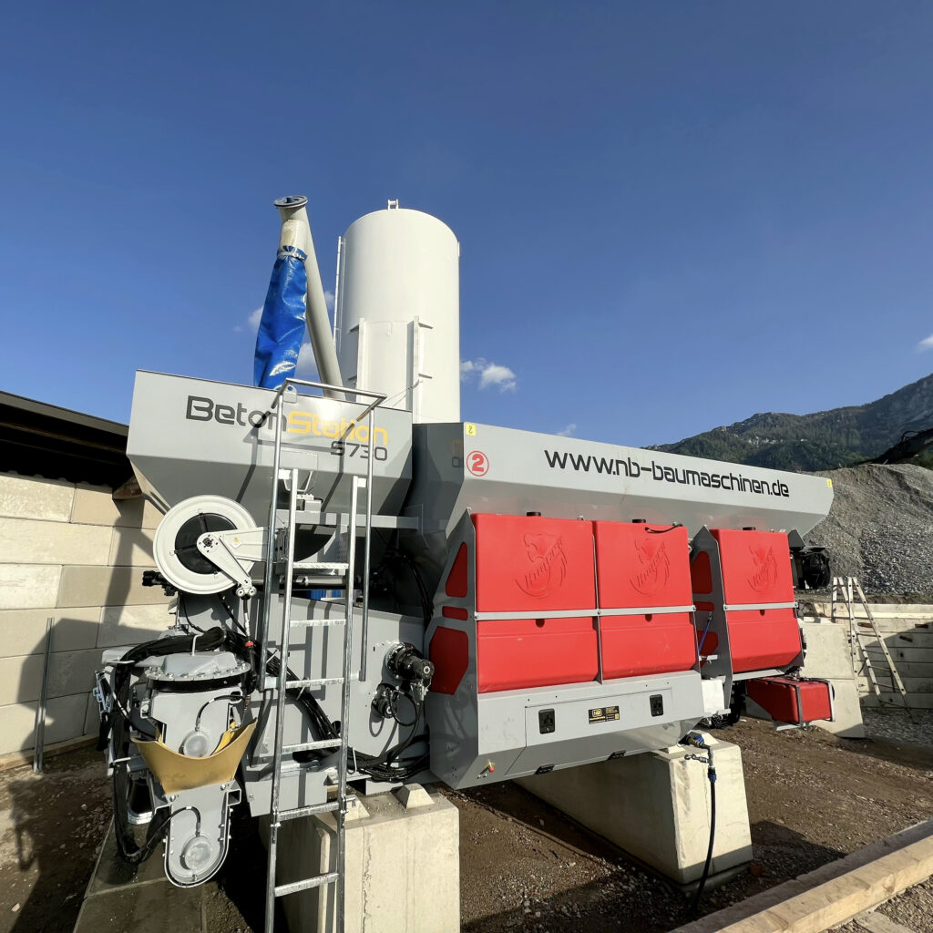 Graue BetonStation S730 von Kimera mit hohem Siloturm und roten Verkleidungen steht vor blauer Bergkulisse unter klarem Himmel.