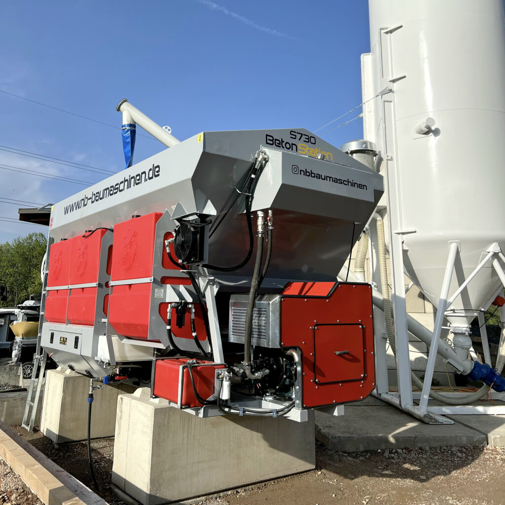 Eine graue und rote BetonStation S730 von Kimera steht neben einem weißen Siloturm unter blauem Himmel, mit Schläuchen, Rohren und Technikmodulen.