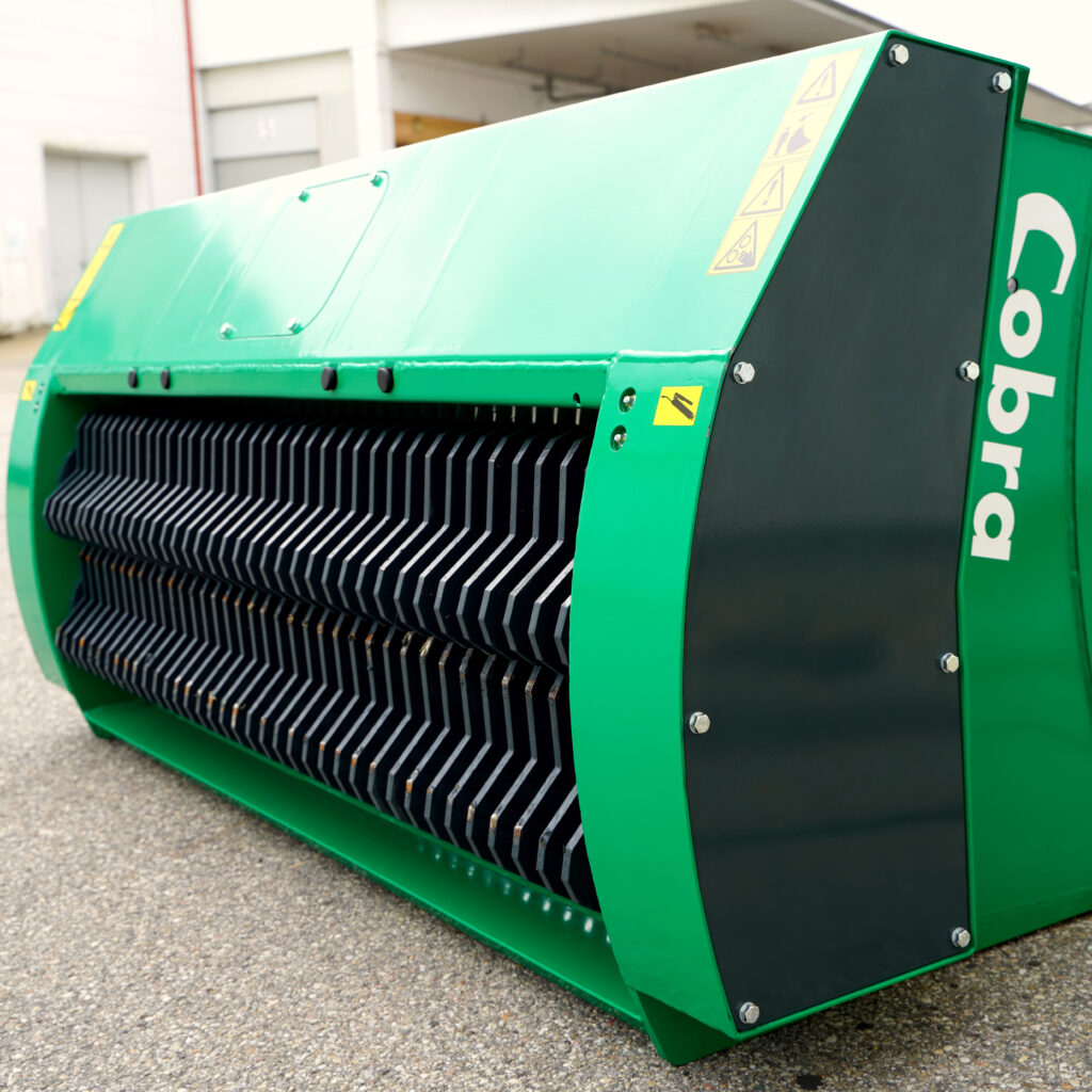 Ein grün-schwarzer COBRA XS2 mit rotierenden Schreddermessern und Warnaufklebern steht schräg vor einem hellen Industriegebäude auf Asphalt.