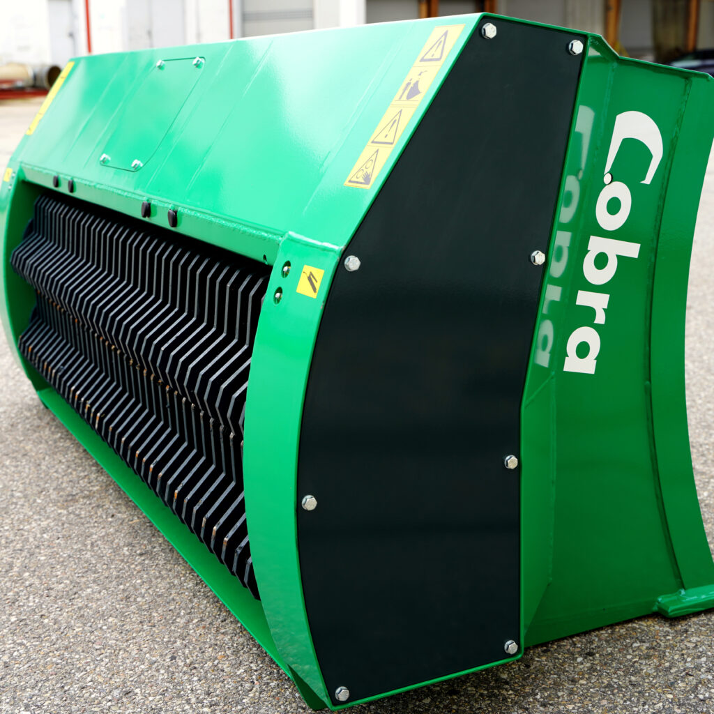 Grüne COBRA-Zerkleinerereinheit mit schwarzem Schutzgitter, Warnsymbolen und bolzenbefestigter Seitenverkleidung in einer Industriehalle.