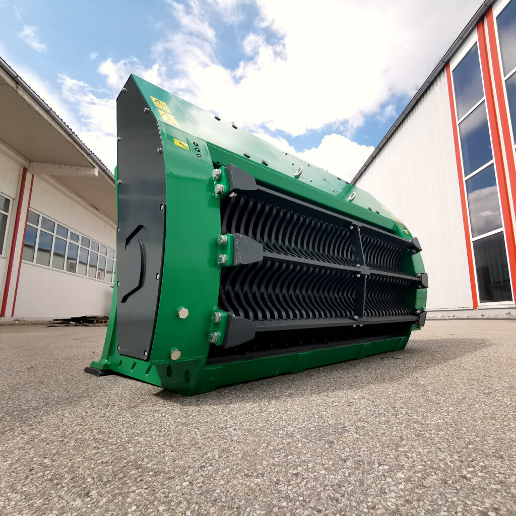 Ein grüner COBRA-S3-Zerkleinerer steht vor Industriegebäuden auf Asphalt, mit schwarzem Schneidwerk und hellem Himmel im Hintergrund.